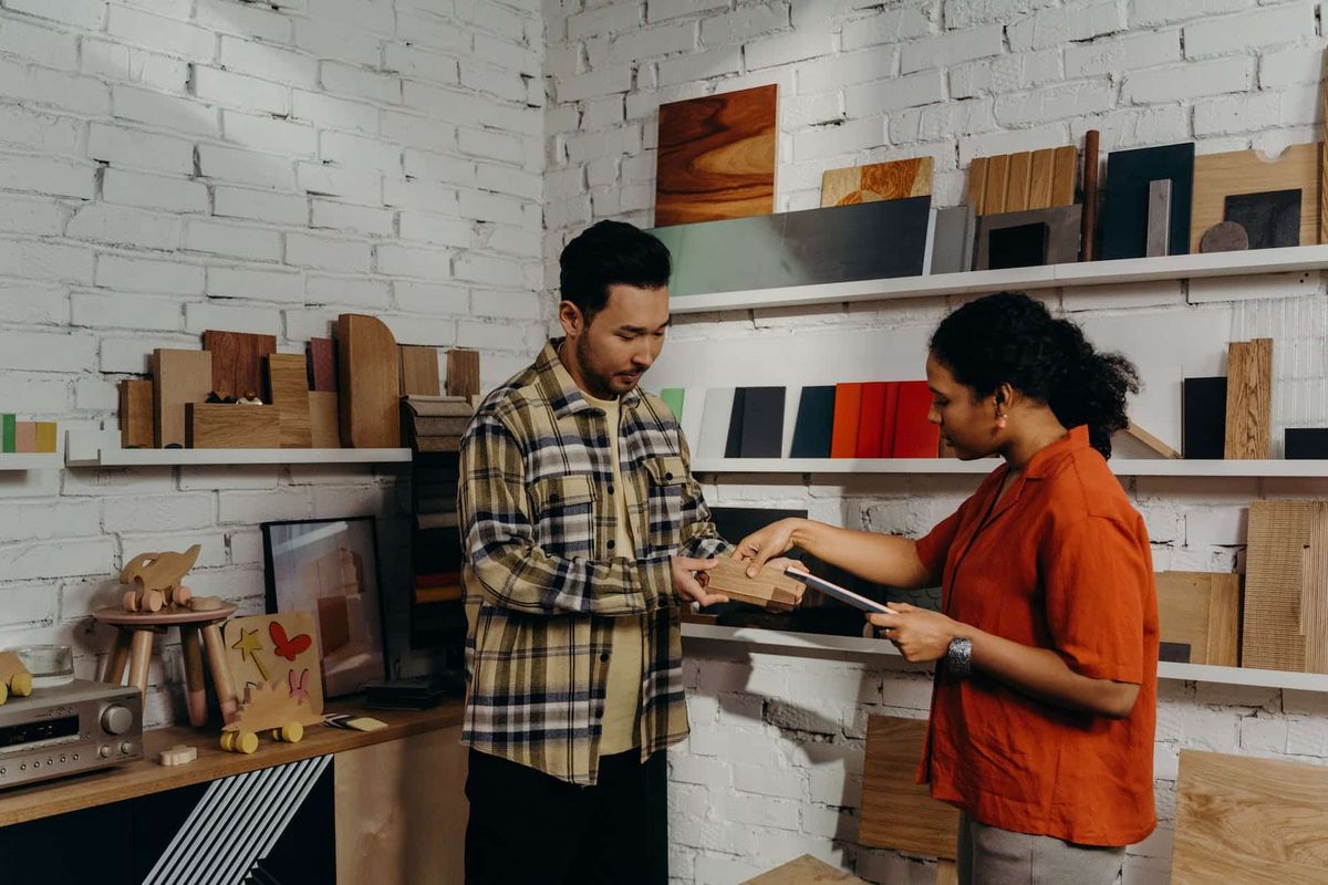 man in plaid button up shirt showing a sample of wooden board to a woman in red shirt